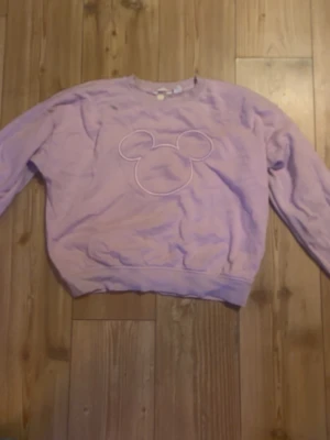 Ljuslila sweatshirt med Disney-tryck XS - Superfin ljuslila sweatshirt från Disney i storlek XS. Tröjan har ett broderat konturmönster av Musse Pigg på bröstet och är gjord i mjuk bomull. Klassisk rund halsringning och långa ärmar. Perfekt för dig som gillar söta och bekväma plagg.