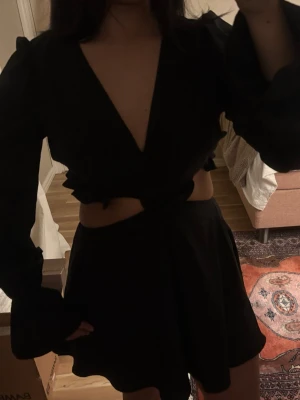 Svart klänning  - Säljer en svart  klänning med långa, utsvängda ärmar och djup v-ringning. Klänning har en cut-out detalj i midjan och knyt framtill. Materialet känns lätt och flowy, perfekt för dig som vill ha något stilrent men ändå edgy.