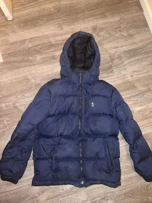Blå pufferjacka från Polo Ralph Lauren - Säljer en mörkblå pufferjacka från Polo Ralph Lauren med huva och dragkedja framtill. Jackan har två sidofickor och en bröstficka med dragkedja. Fylld med 75% dun och 25% fjäder för extra värme. Perfekt för kalla dagar. Jackan har skavts på Ralph lauren märket. dragkedjan har fastnat på fickan men går att lösa för den som vill 
