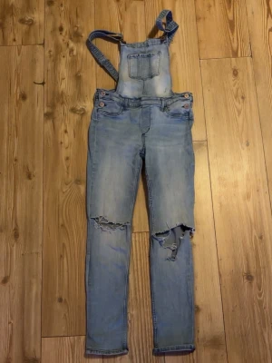 Ljusblå denim hängselbyxor &Denim - Säljer ett par ljusblå hängselbyxor i denim från &Denim. Byxorna har slitna detaljer och hål på båda knäna för en cool, avslappnad look. Klassisk ficka på bröstet och justerbara hängslen. Perfekta för dig som gillar streetstyle.