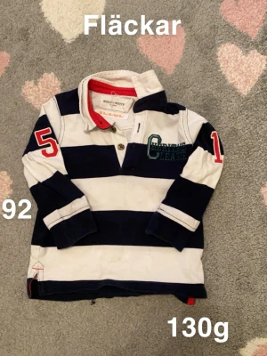 Randig långärmad tröja Holly & White - Säljer en långärmad tröja från Holly & White i storlek 92 soprtig look. Tröjan har breda marinblå och vita ränder, broderad text på bröstet och röda siffror på ärmarna. Kragen är vit med röd insida och knappar framtill.🌸 Fläckar, storlek 92