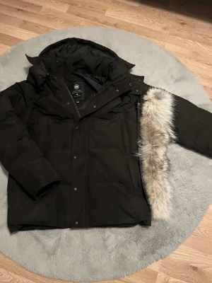 Svart dunjacka från Canada Goose - Säljer en svart dunjacka från Canada Goose med avtagbar pälskant på huvan. Jackan har en klassisk patch på ärmen, dragkedja och knappar framtill samt stora fickor. Perfekt för kalla vinterdagar och riktigt varm. Stilsäker och tidlös modell.