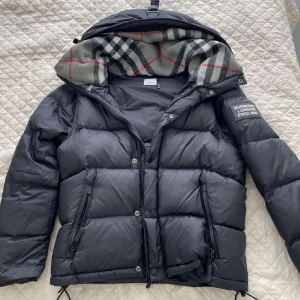 Svart pufferjacka från Burberry - Säljer en svart pufferjacka från Burberry med avtagbara armar om man vill ha den som en väst också. Såklart äkta och inte mycket tecken på användning. Dragkedja lite sönder men går att fixa med limm enkelt. Storlek L men sitter lite mindre som M. (Inga returer)