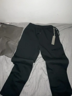 Svarta mjukisbyxor Essentials Fear of God - Svarta mjukisbyxor från Essentials Fear of God med broderad logga på benet och lapp framtill. Byxorna har dragsko i midjan och är gjorda i mjukt bomullsmaterial. Perfekta för en chill och avslappnad stil.