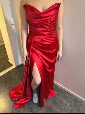Röd axelbandslös långklänning - Snygg röd axelbandslös långklänning i glansigt satinmaterial med draperad överdel och hög slits framtill. Klänningen har en elegant och figurnära passform som ger en lyxig känsla. Perfekt för dig som vill sticka ut med en dramatisk look.