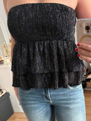 Glittrig svart bandeau tubtopp - Säljer en glittrig svart bandeau tubtopp med volangdetaljer. Ett perfekt festplagg som går att använda både som topp och kjol🤩Endast använd en gång!