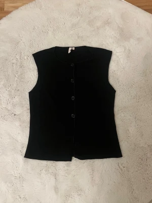 Svart ärmlös topp från Nelly XS - Svart ärmlös topp från Nelly i storlek XS. Toppen har en rak passform och knäpps med fyra knappar framtill. Enkel och stilren design som passar till många olika looks. Oanvänd