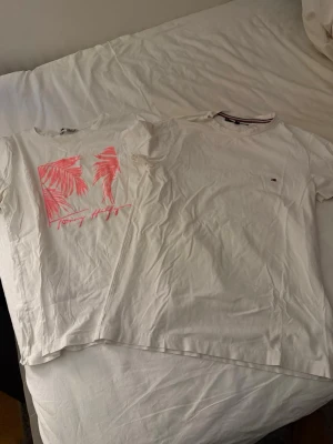 Tommy Hilfiger vita t-shirts - Två vita t-shirts från Tommy Hilfiger. Ena har ett rosa palmtryck och logga framtill, den andra har en liten broderad logga på bröstet. Båda är klassiskt kortärmade och gjorda i mjuk bomull. Säljer dessa tillsammans så priset är för båda, men om man vill ha ena så hör av er!💕