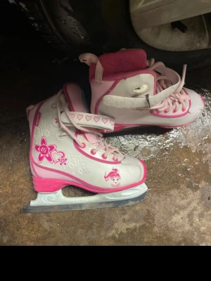 Rosa och vita skridskor från CCM - Säljer ett par skridskor från CCM i vitt och rosa med blommiga och hjärtformade detaljer. Skorna har snörning och kardborreband med hjärtmönster. Utsidan är dekorerad med blommor och en söt figur, perfekt för dig som gillar färgglada och lekfulla detaljer. i behov av att slipas