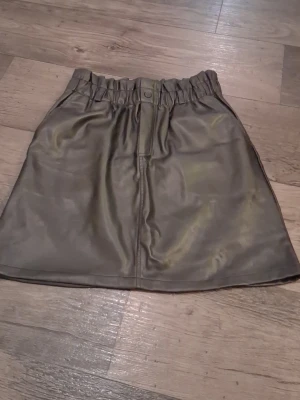 Svart kortkjol i skinn från Noisy May - Säljer en svart kortkjol från Noisy May i snyggt skinnmaterial. Kjolen har resår i midjan, knapp framtill och två bakfickor. Perfekt för dig som gillar en cool och modern stil. Passar dig som vill ha en kjol med lite edge.