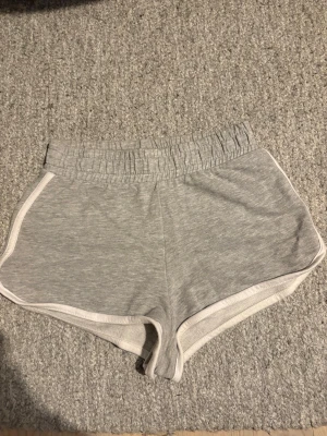 Grå mjuka shorts med resårmidja - Grå shorts i mjukt bomullsmaterial med elastisk midja och vita kanter i storlek XS (passar S) Klassisk sportig look med loose fit och korta ben, perfekta för sommardagar eller chill hemma. Inga fickor eller synliga loggor.