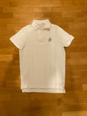 Abercrombie & Fitch polo - Klassisk vit pikétröja från Abercrombie & Fitch i storlek M. Tröjan har korta ärmar, krage och knappar framtill. På bröstet finns en grå broderad A&F-logga. Tillverkad i bomull för en skön och luftig känsla.