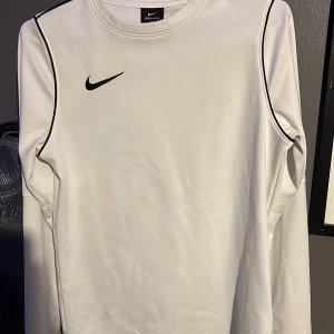 Träningströja  - Säljer en vit långärmad Nike Dri-Fit tröja med svarta detaljer längs axlarna och klassisk Nike-logga på bröstet. Tröjan är tillverkad i ett lätt och funktionellt material som andas, perfekt för träning eller sportiga outfits. Storlek 147-158 men sitter som 160-170