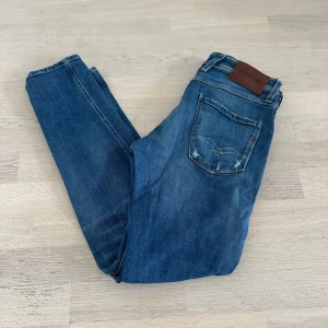 Blå Replay anbass med slitningar  - Snygga blå jeans från Replay med coola slitningar och klassisk femficksmodell. Jeansen har normal passform och raka ben, med detaljer som Replay-läderpatch bak och diskreta slitningar framtill. Perfekta för en avslappnad och trendig look.