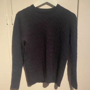 Mörkblå stickad tröja Lexington  - Snygg mörkblå stickad tröja från Lexington i storlek M. Tröjan har ett klassiskt kabelstickat mönster och rund hals. Perfekt för dig som gillar stilrena och tidlösa plagg med lite preppy känsla. Långa ärmar och mjukt material. Det är M men passar S/XS