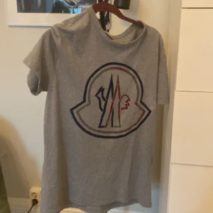 Grå Moncler t-shirt med stor logga - Snygg grå t-shirt från Moncler med stor ikonisk logga i blått och rött framtill med en snygg design. Klassisk passform med rund hals och korta ärmar. Tillverkad i mjuk bomull som är skön mot huden. Perfekt för dig som gillar streetwear och vill sticka ut med ett exklusivt plagg.