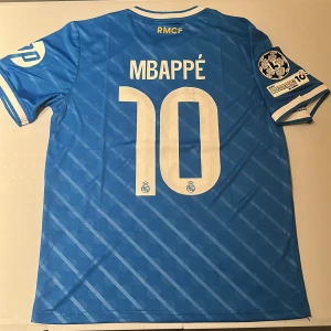 Real Madrid Mbappé 10 blå matchtröja - Blå Real Madrid fotbollströja med Mbappé och nummer 10 på ryggen. Adidas-logga, klubbmärke och FIFA-märke framtill, samt Champions League 15-titlar badge och UEFA Foundation-märke på ärmen. Tunt, ventilerande material och vita detaljer på krage och ärmslut. Replika, endast handtvätt gäller!