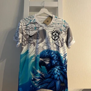Nike Brasil CBF blå/vit fotbollströja S - Unik fotbollströja från Nike med Brasilien CBF-logga. Tröjan har ett stort blått papegojmotiv, grå och vita detaljer samt inslag av turkos. Tillverkad i Dri-FIT-material som andas. Kortärmad och perfekt för dig som vill sticka ut på planen.