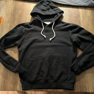 Svart hoodie från Pier One - Svart hoodie från Pier One med klassisk känguruficka och vita snören i huvan. Tröjan har lång ärm och ribbade muddar vid ärmslut och nederkant. Perfekt för en chill och avslappnad stil.