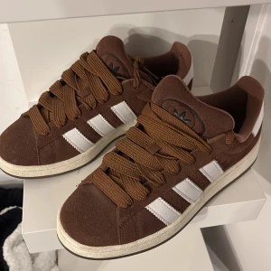 Bruna Adidas Campus sneakers mocka - Snygga bruna Adidas Campus sneakers i mocka med klassiska vita ränder på sidorna och breda bruna skosnören. Skorna har rund tå och platt sula, perfekt för en avslappnad streetstil. Adidas logga på plösen och vit kant runt sulan. Kommer göra de extra rena vid köp