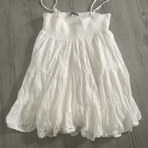 Vit somrig kortklänning med smock - Supersöt vit kortklänning med smock upptill och tunna axelband. Klänningen har volanger och är gjord i ett lätt, luftigt material. Den har en flowy passform och syns inte igenom 
