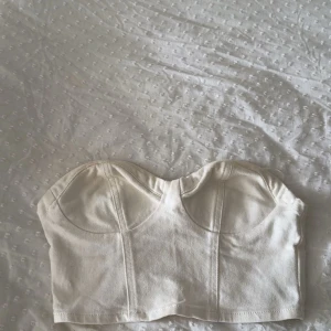 Vit korsett-topp från Perfect Jeans - Säljer en snygg vit korsett-topp från Perfect Jeans i jeansmaterial. Toppen är axelbandslös med markerade kupor och croppad passform. Perfekt att styla med höga byxor eller kjol för en clean look.