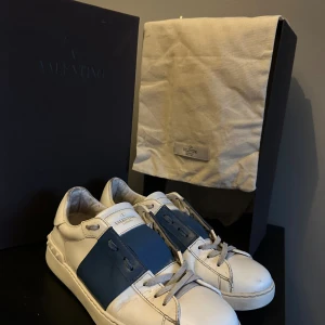 Valentino Garavani Open sneakers vit/blå - Snygga Valentino Garavani Open sneakers i vitt skinn med bred blå panel över vristen. Klassisk rund tå, platt sula och coola nitar på hälen. Skorna har snörning och kommer med originalkartong och dustbag. Perfekta för dig som gillar exklusiva och stilrena sneakers. Storlek 42 1/2 men sitter ca en storlek stort. Box och dustbag medkommer. Pris inte fast i sten, kom gärna med prisförslag. Riktigt fint skick, priset beror på defekten. Vid fler frågor är det bara att kontakta🤩
