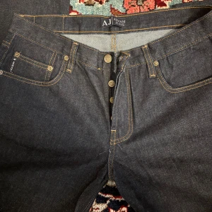 Mörkblå jeans från Armani Jeans, strl 34 - Snygga mörkblå jeans från Armani Jeans med klassisk femficksdesign, kontrastsömmar och silverfärgade knappar. Bakfickan har den ikoniska Armani-loggan. Jeansen är tillverkade i robust denim och har en rak passform.