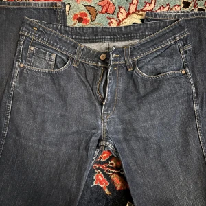 Blå Dylan Raw Denim jeans straight fit - Snygga blå jeans från Dylan i modellen Raw Denim med regular fit och straight leg. Klassisk femficksdesign, kontrastsömmar och knappgylf. Perfekt för dig som gillar en enkel och tidlös look. Materialet är slitstarkt jeans och färgen är mörkblå.