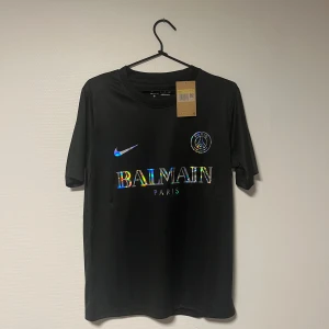 Svart PSG x Balmain fotbollströja Nike S - Svart fotbollströja från Nike i samarbete med PSG och Balmain. Tröjan har korta ärmar, rund hals och är tillverkad i Dri-FIT-material som andas. Framsidan pryds av ett holografiskt Balmain Paris-tryck och PSG-logga. Perfekt för dig som gillar exklusiva samarbeten.