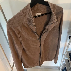 Beige kofta från Murrey med dragkedja - Säljer en beige stickad kofta från Murrey i storlek M. Koftan har en hög krage, dragkedja och knappar framtill. Perfekt för lager-på-lager och chillare dagar. Ribbstickade muddar vid ärmslut och nederkant ger en snygg finish.