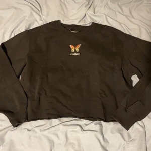 Svart croppad tröja från Hollister - Svart croppad sweatshirt från Hollister med broderad fjäril i orange och gul på bröstet samt Hollister-logga under. Tröjan har rund hals, långa ärmar och ribbade muddar. Perfekt för dig som gillar chill och snygg streetstil.