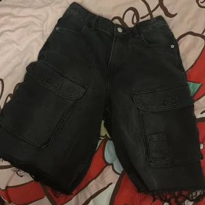 Svarta cargo-jeansshorts med rå kant - Svarta jeansshorts med cargo-stil och stora fickor på sidorna. De har råa, fransiga kanter nedtill för en edgy vibe. Klassisk femficksdesign och knappstängning fram. Perfekta för dig som gillar streetwear och vill ha något som sticker ut.