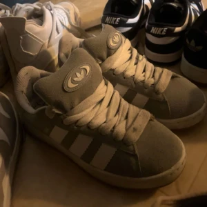 Grå Adidas Campus sneakers i mocka - Säljer ett par grå Adidas Campus sneakers i mjuk mocka med klassiska vita ränder på sidorna och vit sula. Skorna har rund tå, snörning och Adidas logga på plösen och hälen. Perfekta för dig som gillar en clean och stilren look. Storlek 38