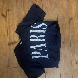Svart croppad topp med PARIS-tryck - Svart croppad topp med stort vitt PARIS-tryck framtill. T-shirten har korta ärmar och en avslappnad passform. Perfekt för dig som gillar streetstyle och vill ha en enkel men snygg statement-piece i garderoben.