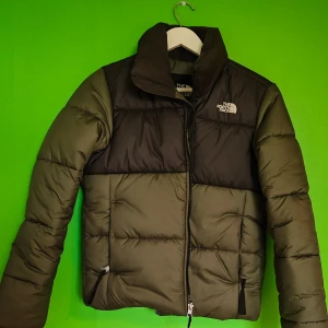Grön och svart pufferjacka The North Face - Säljer en snygg pufferjacka från The North Face i grön och svart färg. Jackan har hög krage, dragkedja framtill och två fickor med dragkedja. Perfekt för kalla dagar och riktigt skön att bära. Broderad logga på bröstet ger en clean look. Knappt använd.