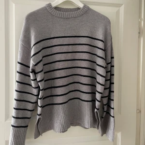 Grå randig stickad tröja - Säljer en grå stickad tröja med svarta horisontella ränder. Tröjan har ribbade muddar och nederkant samt lång ärm. Perfekt för lager-på-lager och chill dagar. Oversized fit med slits i sidorna för extra komfort.