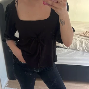 Svart blus med knyt och puffärm - Säljer en svart blus med fyrkantig ringning, puffiga trekvartsärmar och knyt i midjan. Blusen har en lös passform och är tillverkad i ett mjukt, lätt material som faller snyggt. Perfekt att styla med jeans eller kjol.