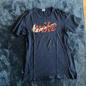 Svart Nike t-shirt med tryck - Svart t-shirt från Nike med stort vintage-inspirerat logotryck i orange och vitt på bröstet. Klassisk rund hals och korta ärmar. Materialet är mjuk bomull som känns skönt mot huden. Perfekt för dig som gillar sportiga och stilrena plagg.