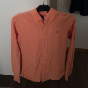 Orange skjorta från Polo Ralph Lauren - Säljer en orange långärmad skjorta från Polo Ralph Lauren i slim fit. Skjortan har klassisk krage, knäppning framtill och den ikoniska gröna loggan broderad på bröstet. Perfekt för dig som gillar stilrena och färgstarka plagg.