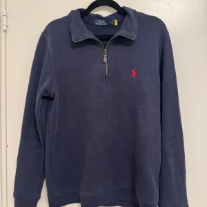 Marinblå half zip tröja Polo Ralph Lauren - Marinblå långärmad tröja från Polo Ralph Lauren med röd broderad logga på bröstet. Tröjan har en half zip-dragkedja vid halsen och är gjord i mjuk bomull. Perfekt för chill dagar och snygg att matcha med jeans eller chinos. Storlek S pris 550 kan diskuteras vid snabbaffär 