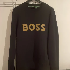 Svart BOSS sweatshirt med guldtryck - Svart sweatshirt från BOSS i storlek S med stort guldigt BOSS-tryck på bröstet. Tröjan har rund halsringning och långa ärmar. Tillverkad i mjukt material.