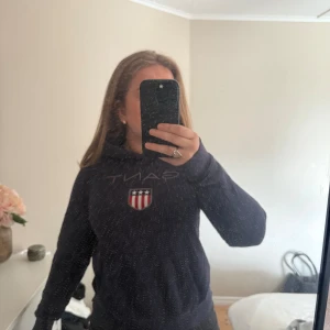 Mörkblå hoodie från Gant med tryck - Mörkblå hoodie från Gant med klassiskt broderat logotyp-tryck och sköld på bröstet. Tröjan har huva, långa ärmar och en relaxed passform. Perfekt för dig som gillar en sportig och stilren look. Bra skick 