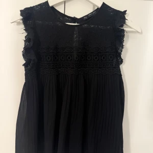 Svart blus från Zara - Supersnygg svart blus från Zara med spets upptill och volangdetaljer vid axlarna. Blusen är ärmlös och har ett plisserat tyg nedtill som ger ett luftigt fall. Perfekt för dig som gillar detaljer och en elegant look.