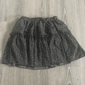 Svart prickig kortkjol med volang - Supersöt svart kortkjol med vita prickar och volangdetaljer. Kjolen har resår i midjan och knytband. Tunt, lätt material som ger en luftig känsla.