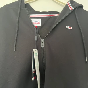 Svart hoodie från Tommy Jeans - Svart hoodie från Tommy Jeans med dragkedja och huva. Snygga detaljer med röd och vit rand på kragen och klassisk logga på bröstet. Tillverkad i mjuk bomull, perfekt för chill dagar. Storlek M. Tag fortfarande kvar aldrig använd. Kontakta för disskution om priset