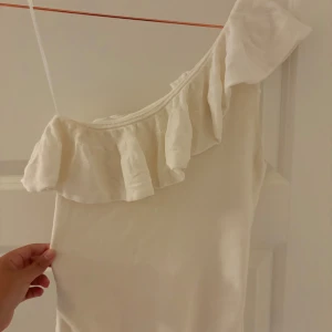 Vit oneshoulder topp med volang från MNG - Säljer en vit oneshoulder topp från MNG i storlek M. Toppen har en snygg volang längs halsringningen och är tillverkad i mjukt bomullsmaterial. Perfekt för dig som gillar en clean och feminin stil med lite extra edge tack vare den asymmetriska axeln.