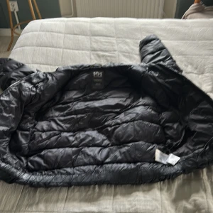 Svart pufferjacka Helly Hansen 164/14 - Säljer en svart pufferjacka från Helly Hansen i storlek 164/14. Jackan är vadderad och har en klassisk pufferlook med breda kanaler. Materialet är glansigt och jackan har lång ärm och dragkedja. Perfekt för kalla dagar när du vill hålla värmen.