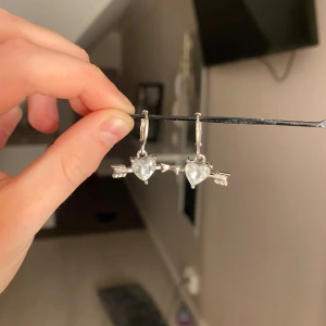 Silverörhängen med hjärta och pil - Säljer ett par snygga örhängen i silverfärgad metall med hängande detaljer formade som hjärtan och pilar. Hjärtat är dekorerat med en klar sten som ger extra bling. Perfekt för dig som gillar unika och romantiska smycken med lite edge. Vet tyvärr inte im dom är nickel.