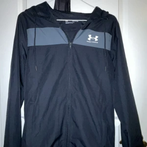 Svart vindjacka från Under Armour XS - Svart vindjacka från Under Armour i loose fit med grå panel över bröstet och vit logga. Jackan har huva med snörning, dragkedja framtill och är tillverkad i lätt polyester. Perfekt för dig som gillar sportig stil och vill ha något smidigt till vardagen. Använd några gånger helt ny inga skador 